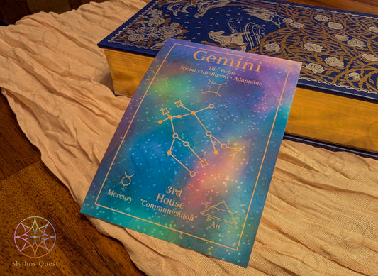 'Gemini' postcard print with astrological design on a tan fabric and wooden textured surface. Propped up on a book with gold trim.