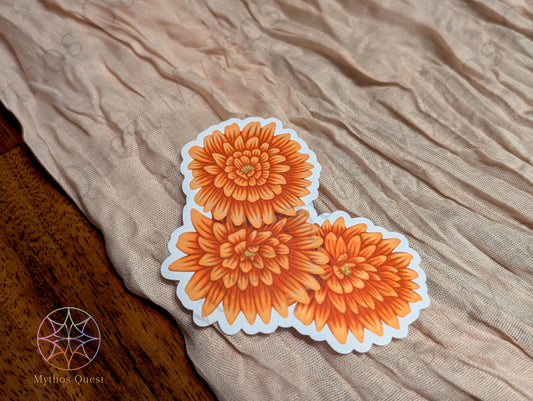 Orange Chrysanthemum (Mum) Sticker laying on top of a tan fabric, resting on top of a wooden surface. Company logo is on the bottom left of the image.